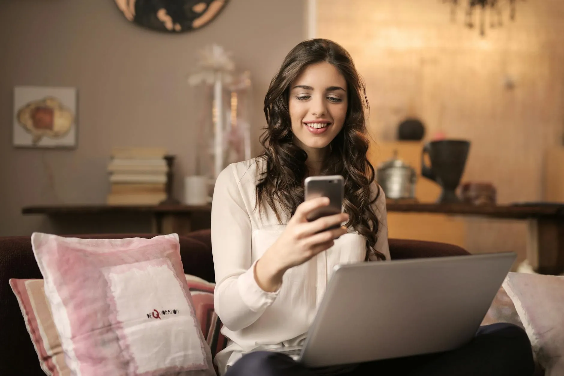 A woman with long brown hair is sitting on a couch, smiling and looking at her smartphone. She has a laptop on her lap and there are decorative pillows beside her. The background shows a warm-toned interior with books and decorative items.