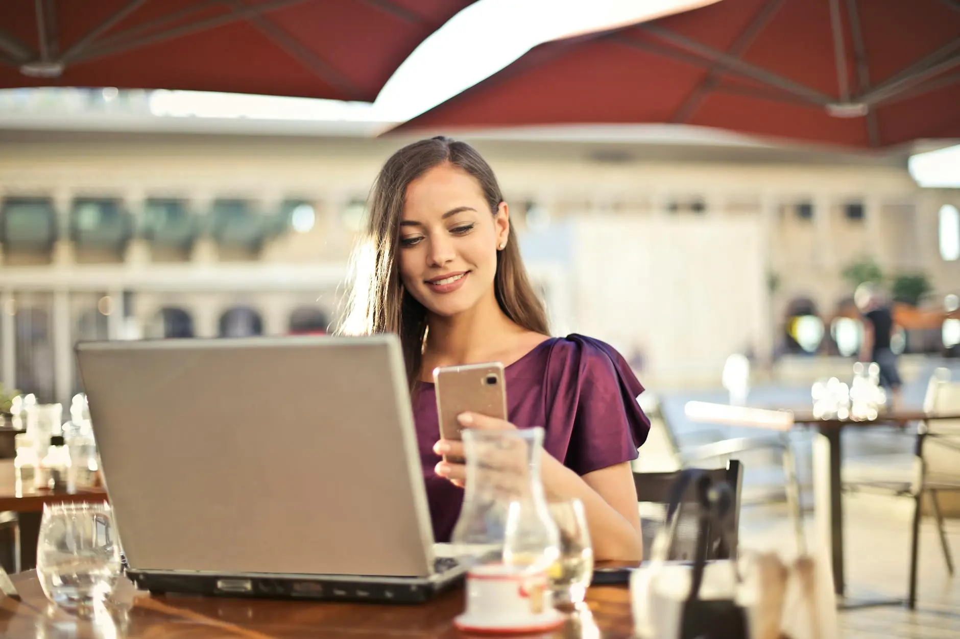 A woman is sitting at an outdoor cafe table using a laptop and looking at her phone. She is wearing a purple blouse, and there are empty glasses on the table. The background shows blurred architecture and large umbrellas.