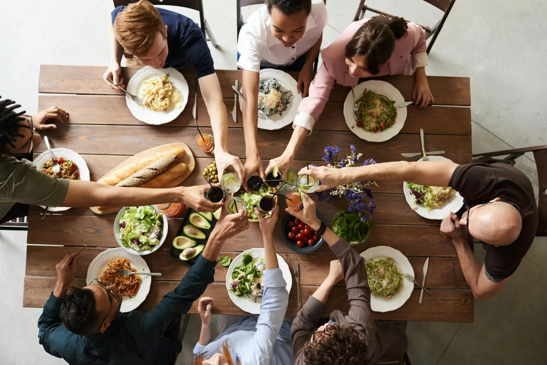 A group of people seated around a wooden table enjoying a meal. They are raising their glasses in a toast. The table is set with various dishes, including pasta, salad, avocados, bread, and drinks.